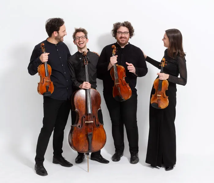 Four musicians in black attire, each holding a string instrument, stand together against a white background, smiling and engaging.