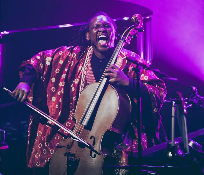 A musician passionately plays a cello on stage, wearing a vibrant patterned robe, with dramatic stage lighting in the background.