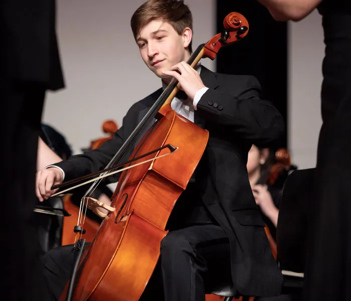 A cellist in a black suit plays the cello during a performance on stage, surrounded by other musicians.