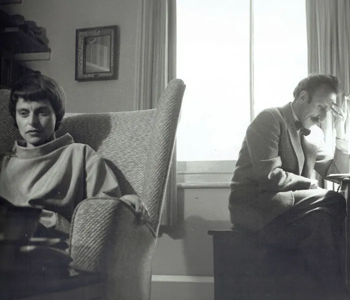 A woman sits in an armchair on the left, looking down, while a man sits on the right by a window, hand on head, appearing deep in thought.