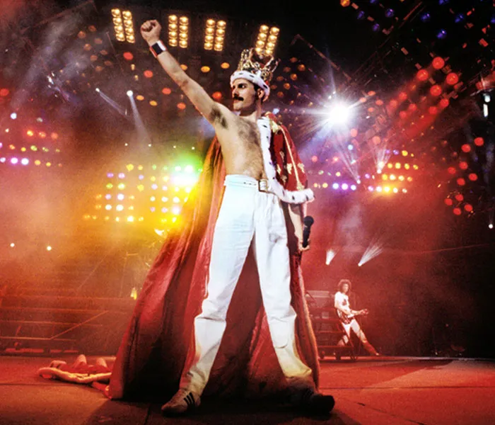 A man wearing a crown and red cape raises his fist on a stage with colorful lights during a performance.