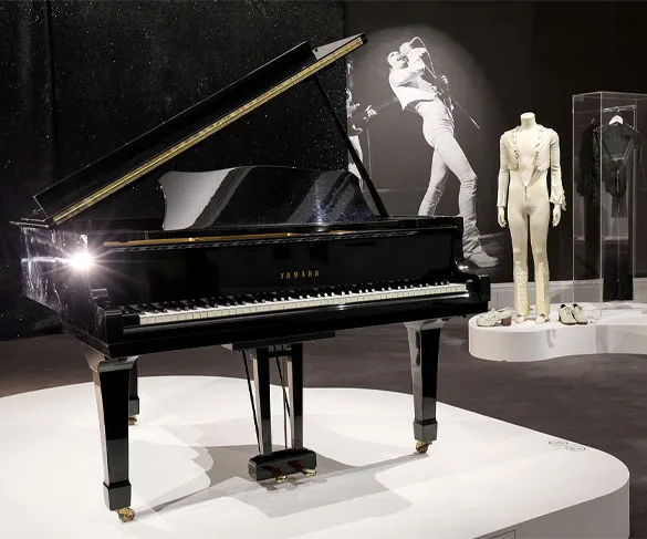 A grand piano is displayed in a museum exhibit with a black-and-white photo and an outfit mannequin in the background.