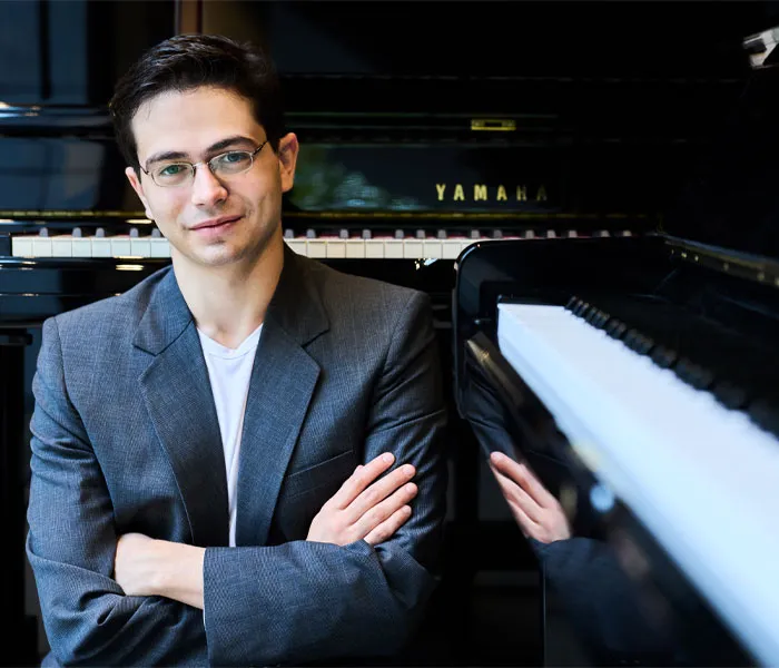 A man in a gray blazer stands with arms crossed in front of a Yamaha piano, smiling at the camera.