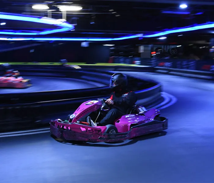 A person wearing a helmet drives a purple go-kart around an indoor track with neon lighting and marked lanes.