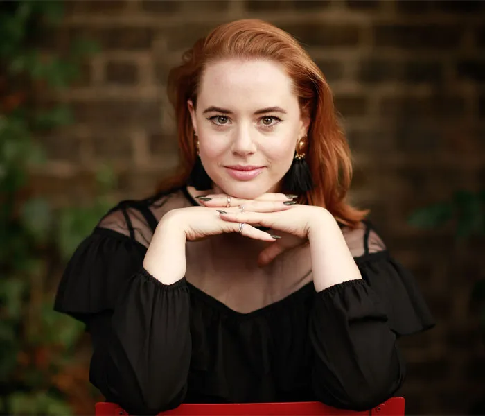 A person with red hair, wearing a black outfit, rests their chin on stacked hands while seated outdoors in front of a blurred brick wall.