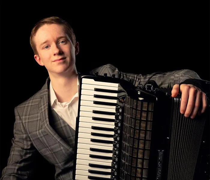 Young person in a plaid suit jacket smiles while holding an accordion against a dark background.