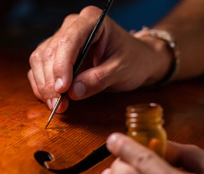 Close-up of a person using a fine paintbrush to apply varnish or paint to a wooden surface, with a small bottle nearby.