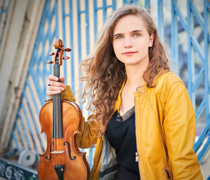 A woman with long, wavy hair in a yellow jacket holds a violin while standing in front of a blue, metal fence.