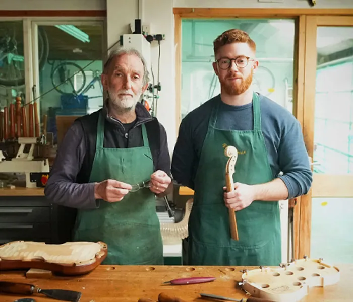 Two men wearing green aprons stand in a woodworking shop, holding woodworking tools and parts of wooden instruments.