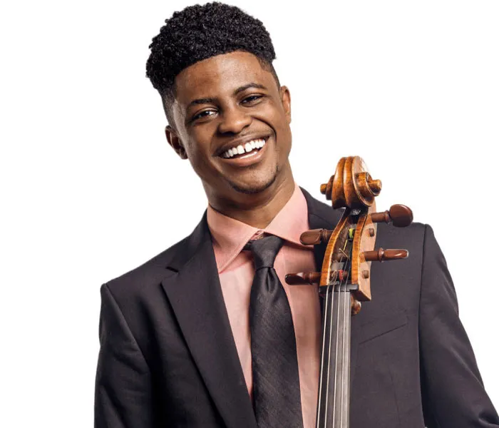 Smiling man in a suit holds a cello, standing against a white background.