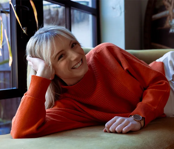 A woman wearing an orange sweater reclines on a sofa, smiling at the camera in a relaxed indoor setting.