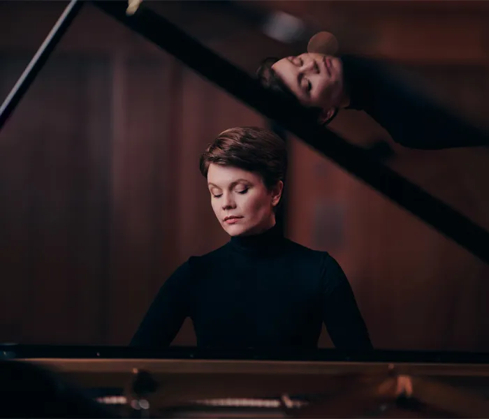 A person with short hair plays a grand piano, with their reflection visible on the open lid of the piano.